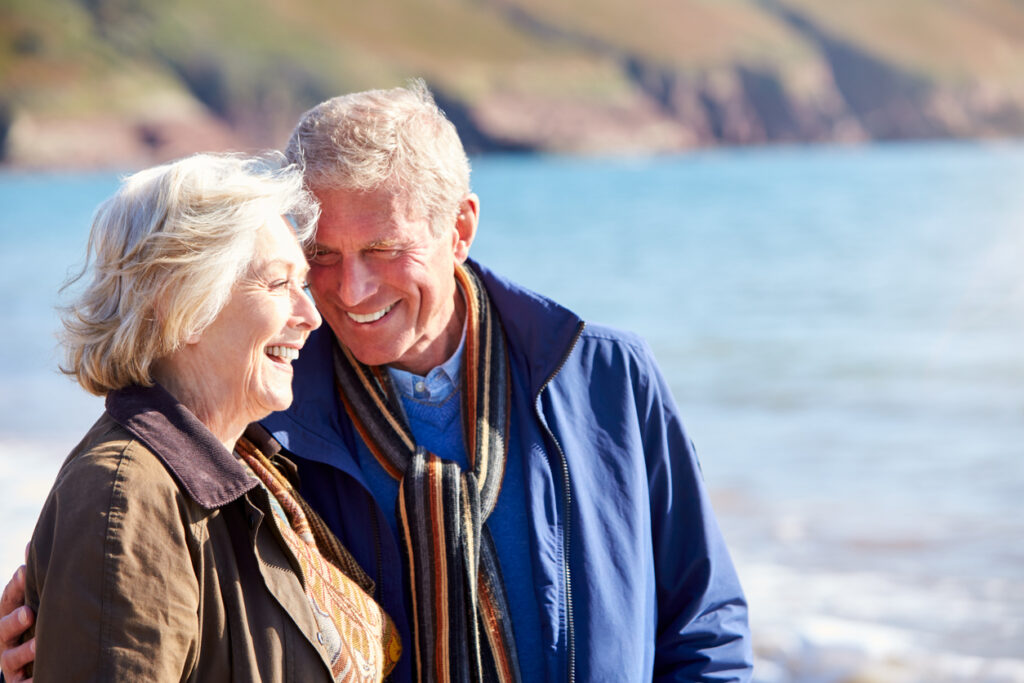 Loving senior couple hugging as they walk along shoreline of beach by waves