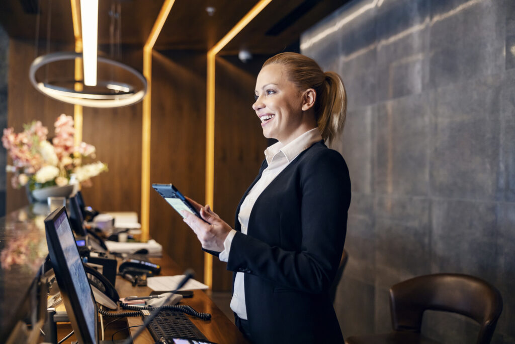 A friendly receptionist is talking to a client while making a reservation on a tablet at hotel reception.