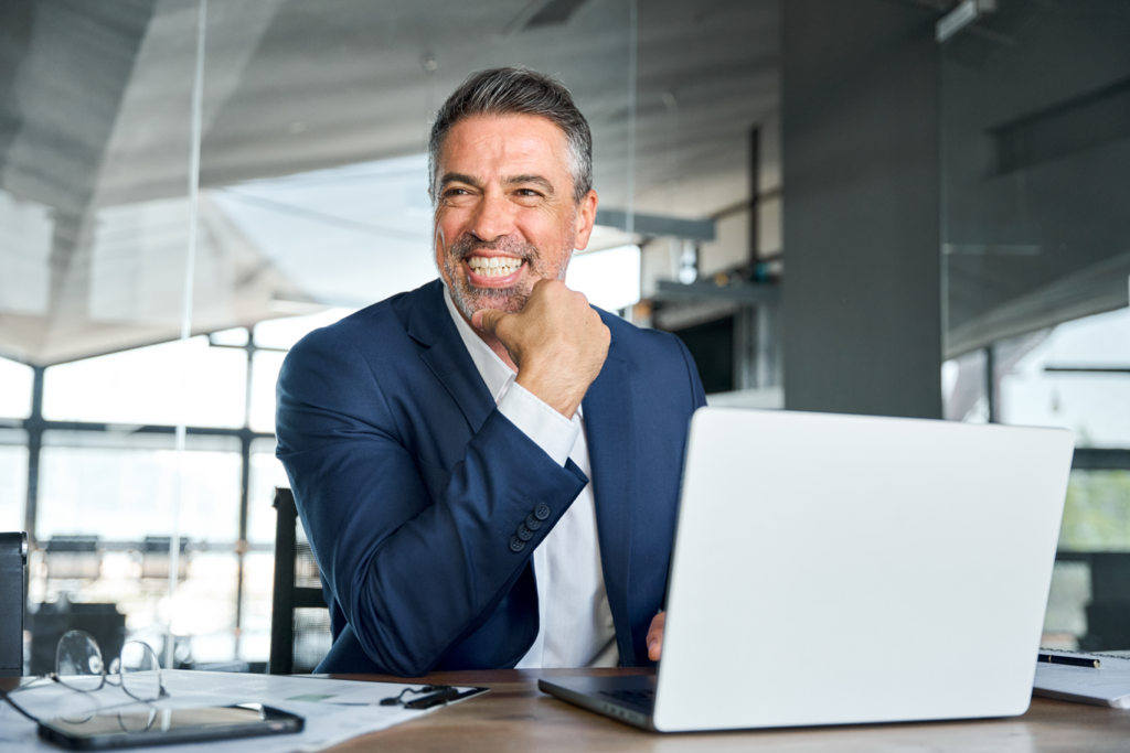 A man in a suit smiles while sitting at a desk with a laptop.